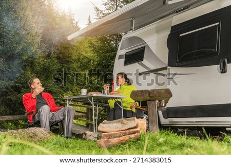 Two people sit at a table by a camper, sharing food and drinks in a forest setting during daylight.