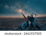 two people sitting in the desert watching the stars and Milky Way next to a telescope, stargazing and exploration concept 