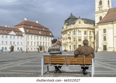 Two People Sitting On A Bench In The Famous Piata Mare, Large Square, In Sibiu, Romania, Rest And Observe Passersby