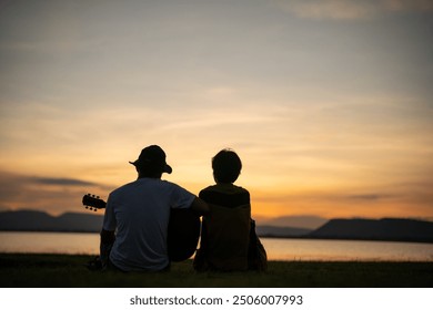 Two people are sitting on the grass by the water, one of them holding a guitar. The sunset in the background creates a peaceful and romantic atmosphere - Powered by Shutterstock