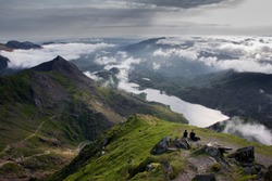 Two People Sitting On Rock At Top Of Mount Snowdon Looking Down Over Dramatic Lakes And Valley