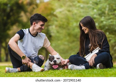 Two People Sitting In A Park With An American Bully Dog Lying Down In The Middle