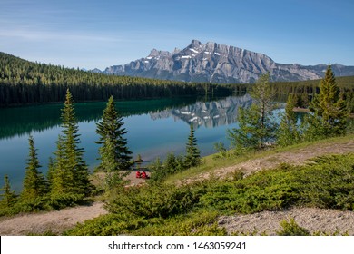 Two People Sitting In Red Adirondack Chairs At Two Jack Lake. Mountain Scenic Reflection Landscape.