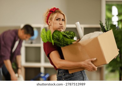 Two people are sorting and moving items in a home. One person holds a box filled with plants and paper while looking serious, and the other looks busy in the background.
