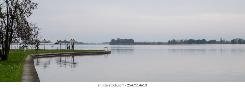 Two people stand by tranquil lakeside huts under overcast skies, evoking a serene autumn ambiance and peacefulness - Powered by Shutterstock