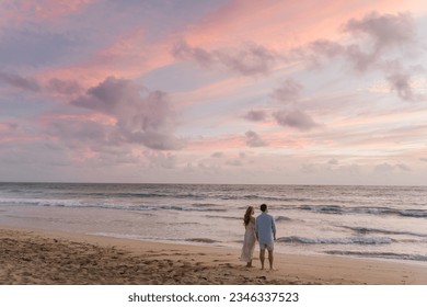 Two people stand on the beach with their backs to the camera, facing the sea during sunset. They gaze into the distance at the pink-hued sunset. The clouds in the sky are also tinged with pink. It's t