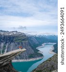 Two people stand on the edge of Trolltunga, a cliff overlooking a scenic fjord in Norway. The sky is clear and the water is turquoise. a couple of men and women hiking the Trolltunga, Norway