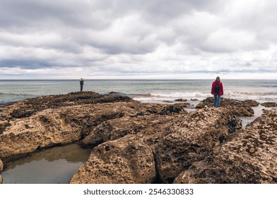 Two people standing on rocky outcrops by the ocean under cloudy skies, enjoying the view of the sea. Coastal scenery with waves, textured rocks, and serene atmosphere. - Powered by Shutterstock