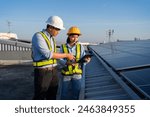 Two people are standing on a roof, one of them is pointing at a tablet. They are wearing safety vests and hard hats. Electrical engineer installs solar panels in power station