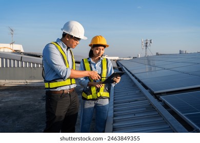 Two people are standing on a roof, one of them is pointing at a tablet. They are wearing safety vests and hard hats. Electrical engineer installs solar panels in power station - Powered by Shutterstock