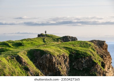 Two people standing at the top and tip of a outcrop or headland. The ground is covered in lush green grass. There are valleys in the ridge with a hiking trail path. The coastline is rugged and rocky. - Powered by Shutterstock