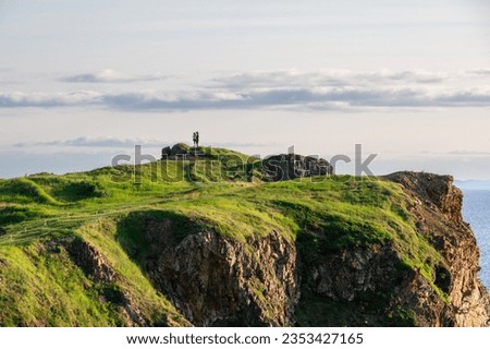 Two people standing at the top and tip of a outcrop or headland. The ground is covered in lush green grass. There are valleys in the ridge with a hiking trail path. The coastline is rugged and rocky.