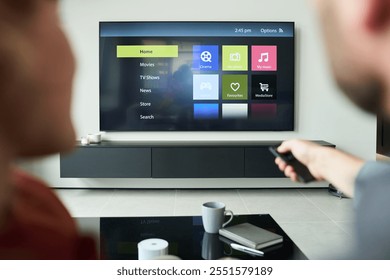 Two people using remote control while seated and facing smart TV displaying various options on screen. Coffee table with mugs and remote control seen in foreground