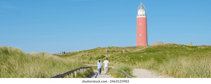 Two people walk down a path next to the lighthouse in Texel, Netherlands, with waves crashing against the shore in the background. a couple at The iconic red lighthouse of Texel Netherlands