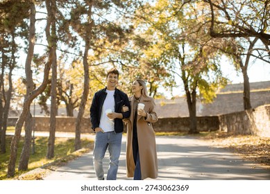 Two people walk down a path in a park. They hold coffee cups and smile at each other. Trees surround the pathway. It is sunny and the leaves are changing color.