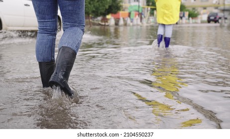 Two People Walk Through Puddles Of Flooded City Rubber Boots On Their Feet, Teamwork, Girls Summer Raincoats, Lot Of Rainwater Is Poured On Street, Natural Weather Phenomena, Walking Boots On Road