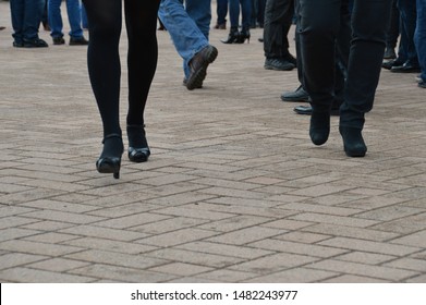 Two People Are Walking Away From A Crowd On City Street. Female In Black Stockings And High Heels With Her Bodyguard Or Friend Leaves A Group Of Men. Concept Of Disagreement Or Completed Transaction.