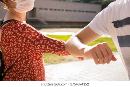 Two people wearing a mask greeting each other with their elbows on the street (with the health center in the background) to maintain the rules of social distancing during the process of new normality 