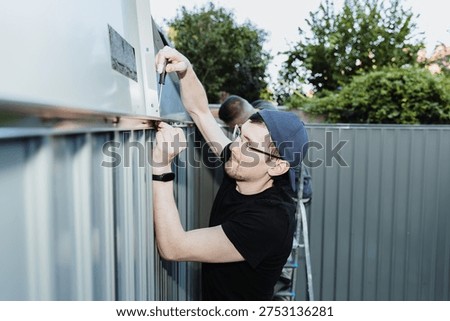 Two people work together to install the roof parts onto the metal shed using screws.