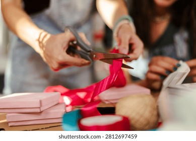 Two people work together to pack gifts. One person uses scissors to cut a red ribbon while the other focuses on the packaging. The scene shows a workspace with materials and supplies.