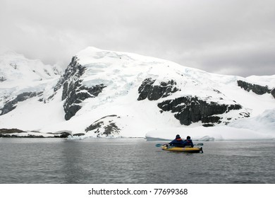 Two People In Yellow Kayak In Antarctica. Shore Under Snow And Cloudy Sky.