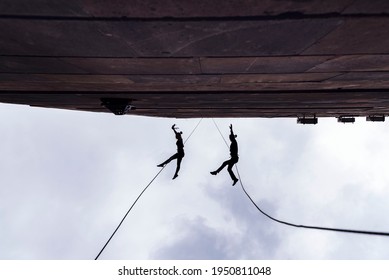 Two Person Silhouettes, Dancing In The Air, Tied By The Ropes On The Roof. Extreme Rope Dance On The Wall Of A Tall Building. 