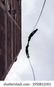 Two Person Silhouettes, Dancing In The Air, Tied By The Ropes On The Roof. Extreme Rope Dance On The Wall Of A Tall Building. 