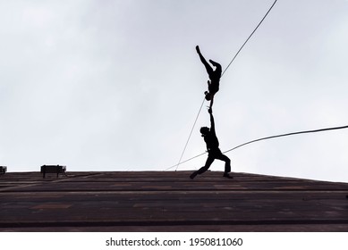 Two Person Silhouettes, Dancing In The Air, Tied By The Ropes On The Roof. Extreme Rope Dance On The Wall Of A Tall Building. 