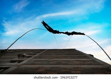 Two Person Silhouettes, Dancing In The Air, Tied By The Ropes On The Roof. Extreme Rope Dance On The Wall Of A Tall Building On The Sunset. 