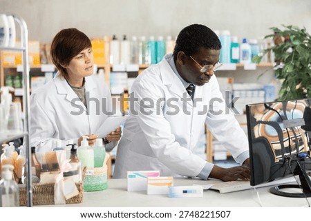 Two pharmacist employee using computer, reads service protocol update, finds out changes, and becomes familiar with work regulations. Woman and African man pharmacists at PC.