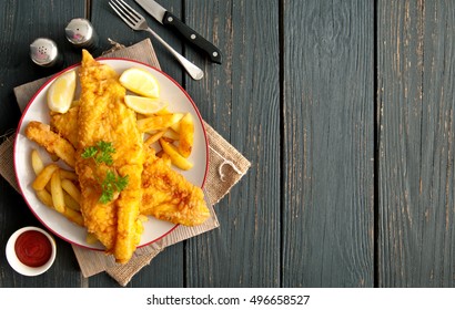 Two pieces of battered fish on a plate with chips on a wooden table with space 