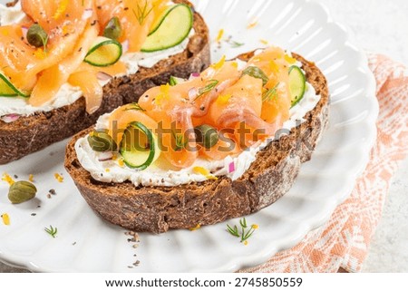 Two pieces of bread with a layer of cream cheese are topped with slices of smoked salmon and cucumber. The dish is placed on a white plate in a kitchen setting.