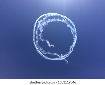 Two Planes Flying In The Sky Making Circle Rings Of Smoke At An Air Show, As A Skydiver Parachutes With An American Flag With A Blue Sky Background