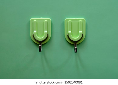 Two plastic adhesive utility wall hangers with metal hooks for hanging clothes, hats, towels seen attached on a green surface background - front, simple, clean closeup view.