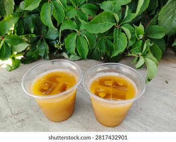 Two plastic cups of orange-colored iced beverage on a light stone surface against lush green foliage, refreshing cold drinks served outdoors in summer