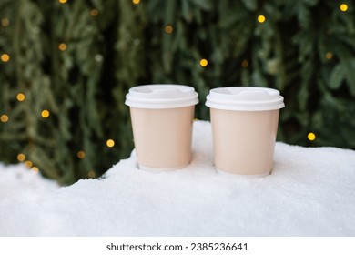 Two plastic cups stand on the snow against the backdrop of a green wall of a Christmas tree and bokeh from a garland. Selective focus, noise. Coffee to go, a walk in the fresh air - Powered by Shutterstock