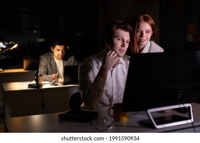 Two Pleasant Business People Working Together On Computer, Discussing Project In Office At Late Night. Side View On Redhead Lady And Handsome Guy Colleagues Brainsotrming. In Dark Boardroom