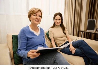 Two Pleasant European Middle-aged Women With A Catalog, Smile Looking At Camera, Sit On Comfortable Sofa In A Modern Cozy Light Interior Of An Office Building. Business, Communication, Interior Design