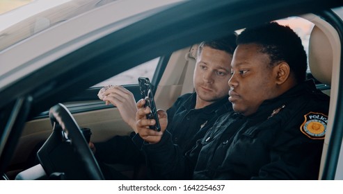 Two Police Officers Best Friends Talking Heart To Heart Sitting Inside Patrol Car During A Break At Work. Handsome Policeman Eating Doughnuts And His Colleague Using A Smartphone.