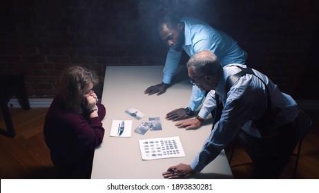 Two police officers and woman criminal in interrogation room. Multiethnic detective showing evidence to female suspect in handcuffs accusing her of crime