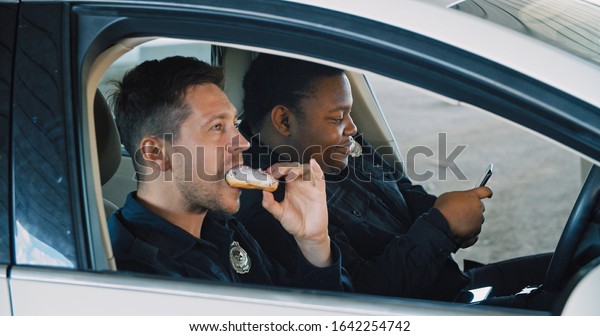 Two policemen on duty
sit inside the police car on urban street. Police officers friends
talking together eating doughnuts and watching videos on smartphone
during break.
