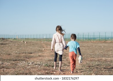 two poor children family brother with toy and thin sister refugee illegal immigrant walking barefooted through hot desert towards state border with barbed fence wire