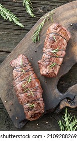 Two portions of juicy freshly roasted beef tenderloin steaks tied up with cooking thread, black pepper and fresh green rosemary stems on wooden cutting board. Natural wood table background. Flat lay.