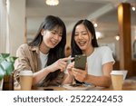 Two positive and charming Asian female friends are looking at something on a smartphone, laughing, and enjoying a conversation while meeting at a coffee shop. people, lifestyles, wireless technology