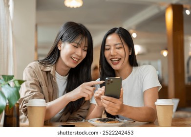 Two positive and charming Asian female friends are looking at something on a smartphone, laughing, and enjoying a conversation while meeting at a coffee shop. people, lifestyles, wireless technology - Powered by Shutterstock