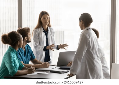 Two positive diverse female doctor colleagues in whites coats discussing healthcare problems, medical job case on professional council, standing at team meeting table, talking - Powered by Shutterstock