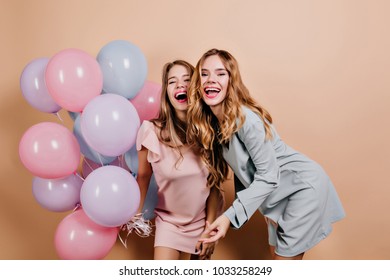 Two positive girls in trendy dresses fooling around together at event. Studio shot of laughing sisters with balloons celebrating holidays.