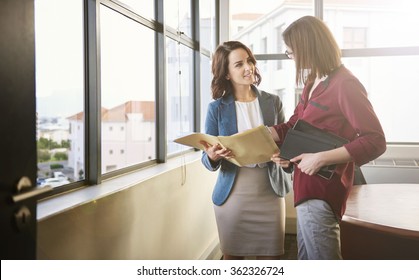 Two positive looking businesswomen standing in an office with large windows looking at each other while discussing some paperwork in a folder with soft sunflare falling through the windows