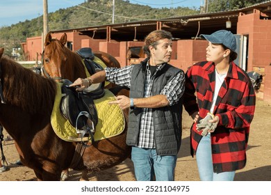 Two positive male female grooms talking and checking saddle on brown horse for riding in yard of stables - Powered by Shutterstock