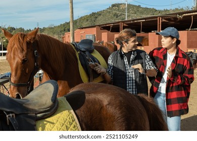 Two positive male female grooms talking and checking saddle on brown horse for riding in yard of stables - Powered by Shutterstock